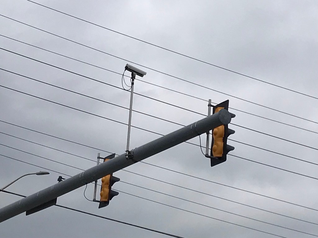 Geometric design of parallel powerlines beyond a diagonally angled bar upon which two traffic lights are mounted along with a surveillance camera aimed downward at the intersection's traffic, all in a closeup view set against an overcast sky