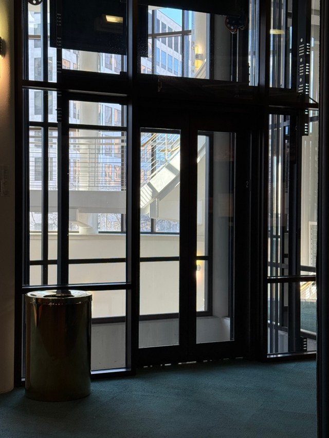 Steel lines of glass elevator shaft looking out into atrium and beyond to office building