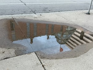 Grohmann Museum, reflected in a puddle on an overcast day