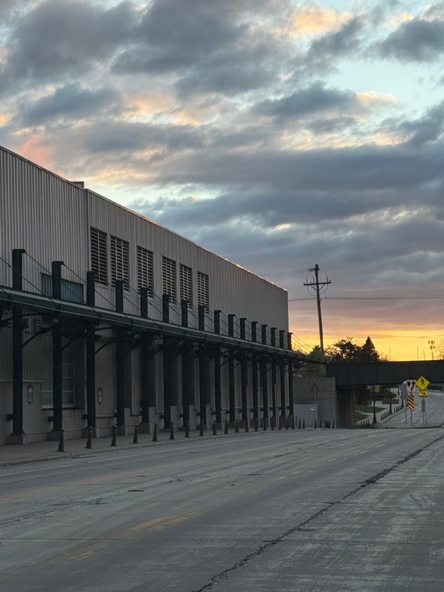 Sunset, Miller Valley, Milwaukee, Wisconsin. A manufacturing building's roofline is limned with setting sunlight, along an empty road, with a railroad bridge overpass and a sky filled with gray fluffy clouds mixed with orange sunlight